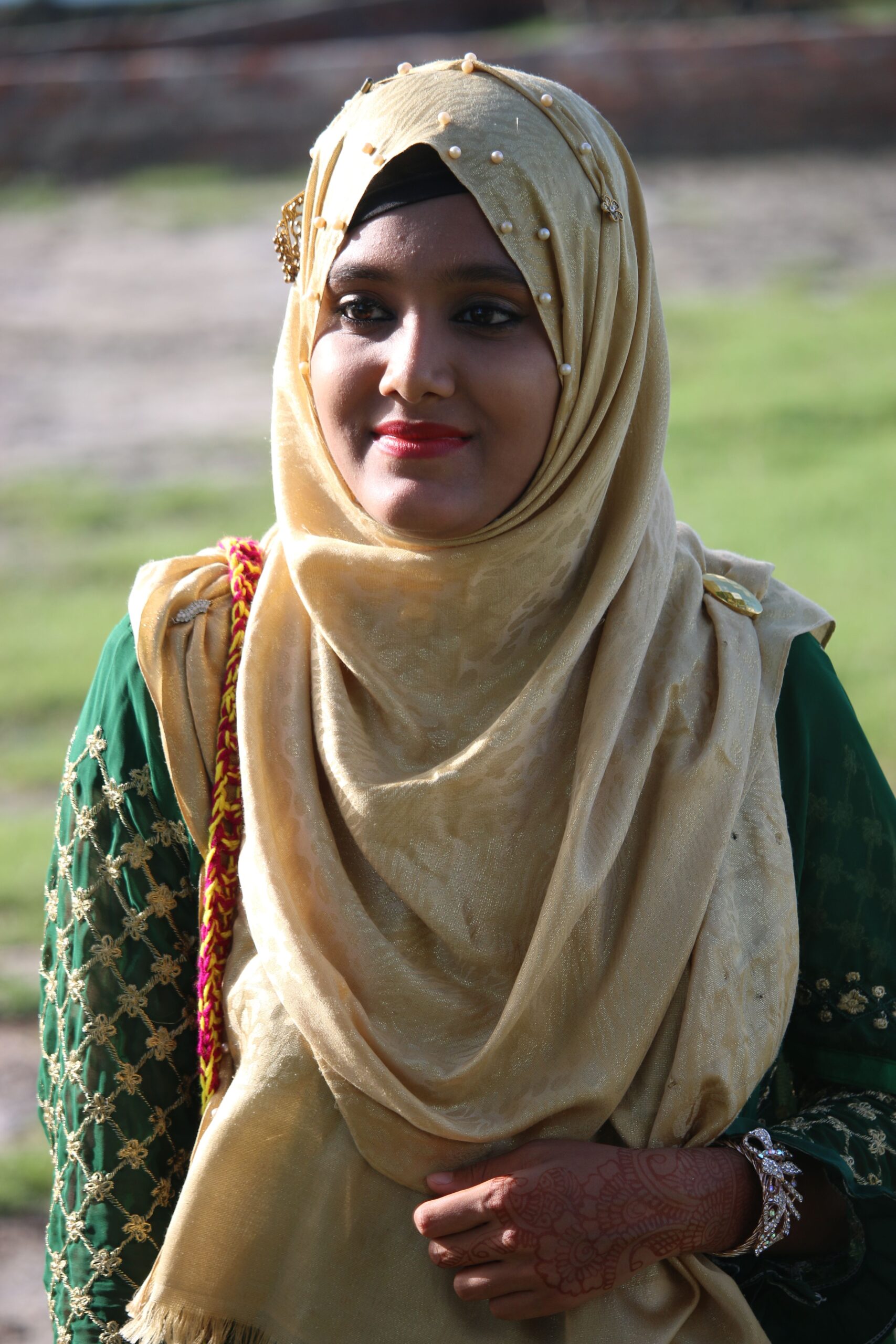 Home A young woman in traditional hijab attire smiles warmly, showcasing cultural fashion outdoors.